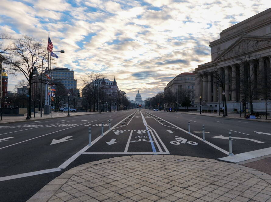 Wide view down Pennsylvania Avenue in Washington, DC, with bike lanes, flags, and the U.S. Capitol in the distance under a cloudy sky.